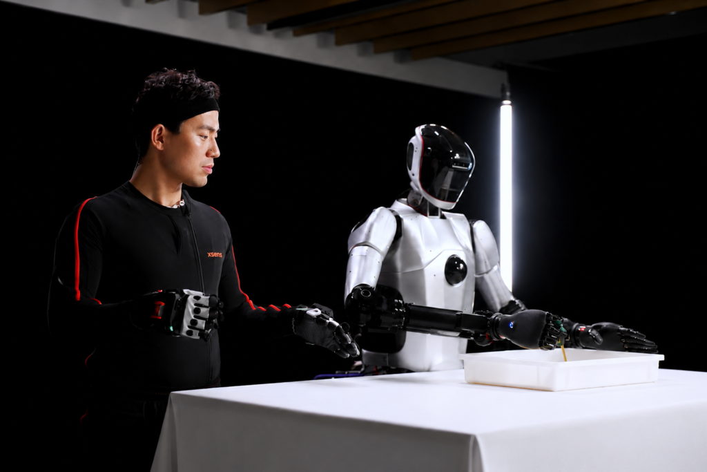 人型ロボットとXsensモーションキャプチャを着用した男性が、テーブル上で手の動作を比較・検証している実験シーン。模倣学習。